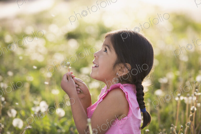 A young child in a field of flowers, blowing the fluffy seeds off a dandelion seedhead…