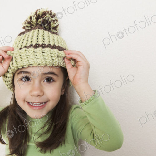 A young child with long brown hair, wearing a knitted hat with a pompom, peering from…