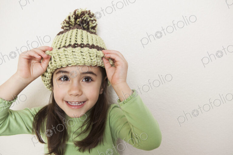 A young child with long brown hair, wearing a knitted hat with a pompom, peering from…