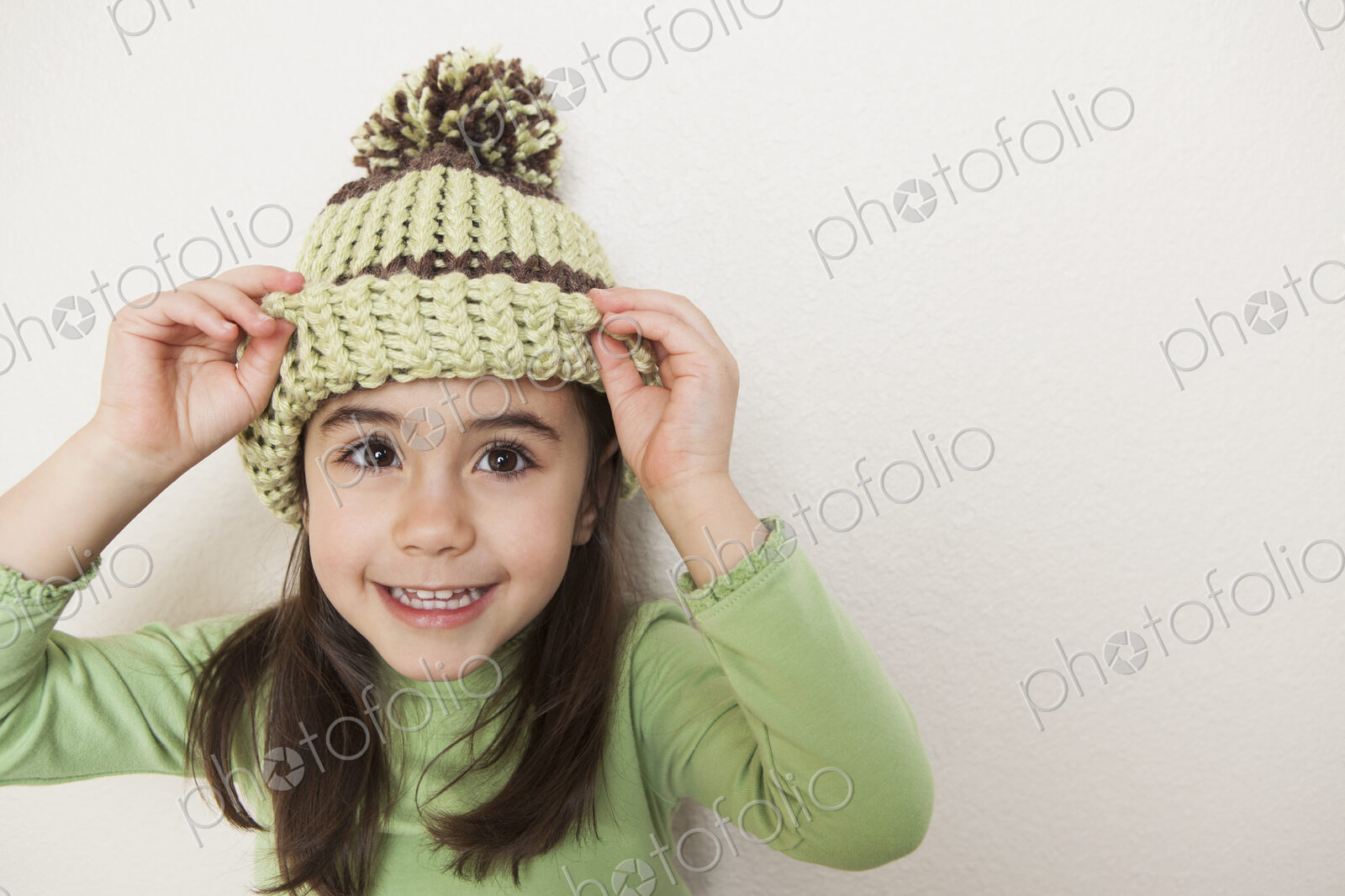 A young child with long brown hair, wearing a knitted hat with a pompom, peering from…