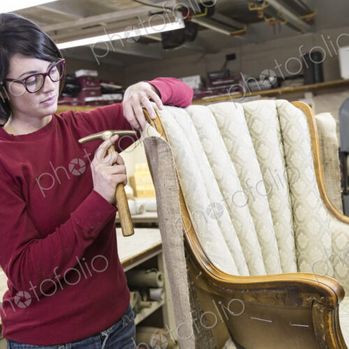 A young female upholsterer using a tack hammer on a chair in an upholstery shop. A young female upholsterer using a tack hammer on a chair in an upholstery shop.