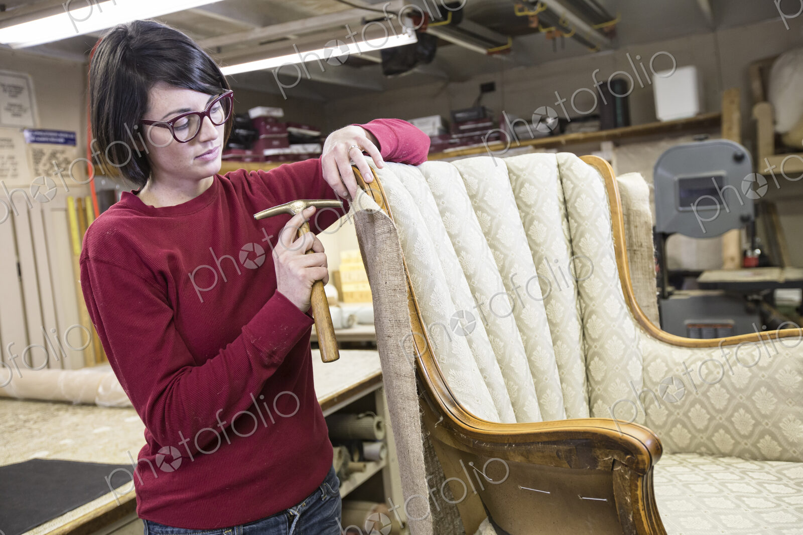 A young female upholsterer using a tack hammer on a chair in an upholstery shop.