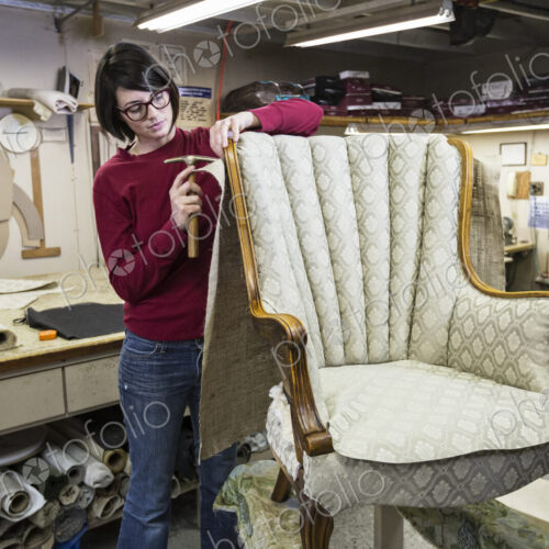 A young female upholsterer using a tack hammer on a chair in an upholstery shop.