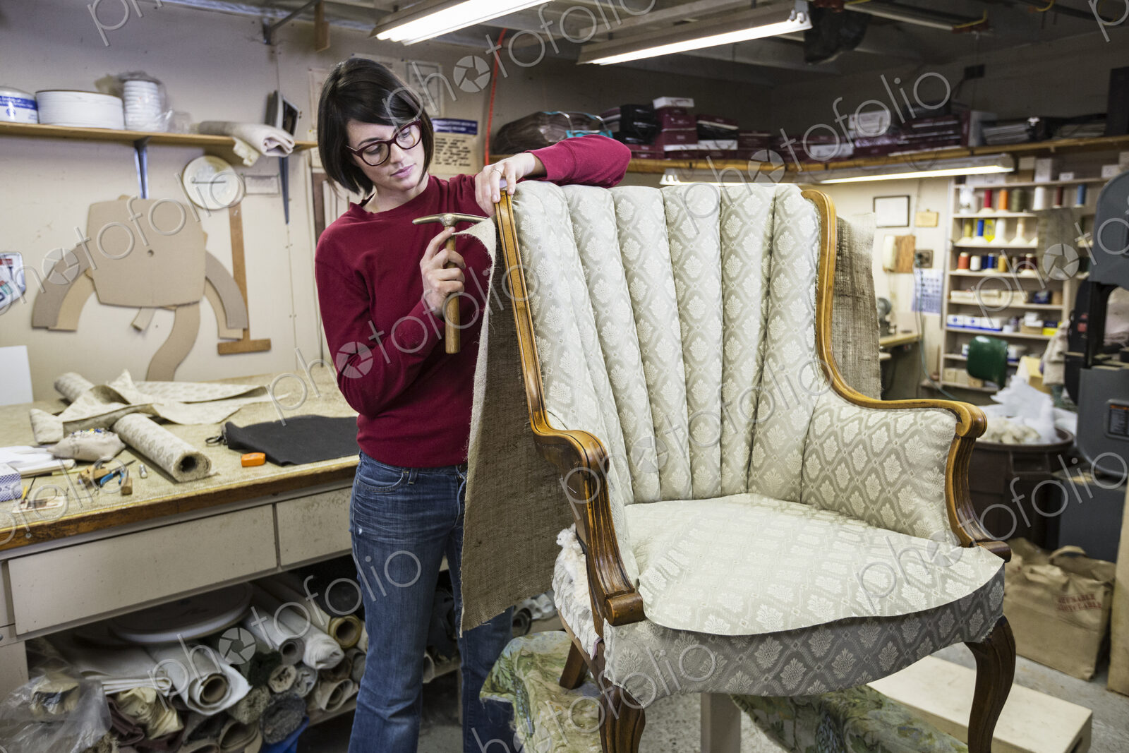 A young female upholsterer using a tack hammer on a chair in an upholstery shop.