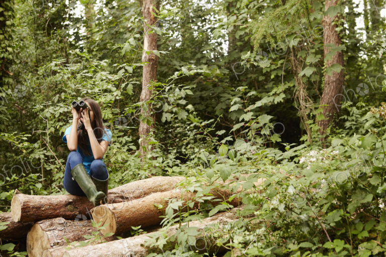 A young girl, a birdwatcher sitting on a log in the woods, using binoculars.