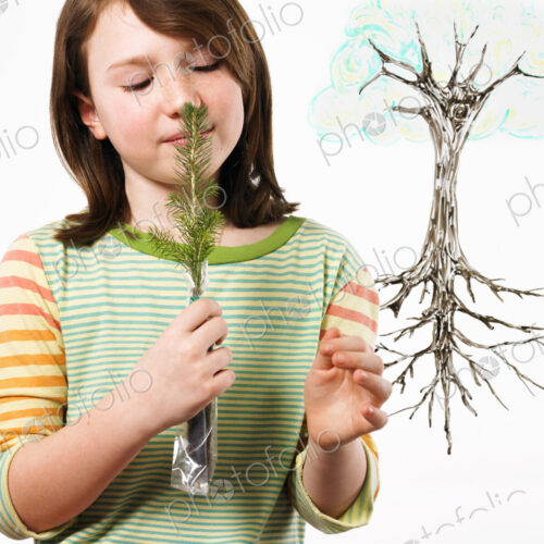 A young girl holding a small evergreen seedling to her nose and smelling it. An…