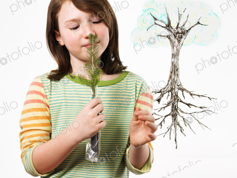 A young girl holding a small evergreen seedling to her nose and smelling it. An…