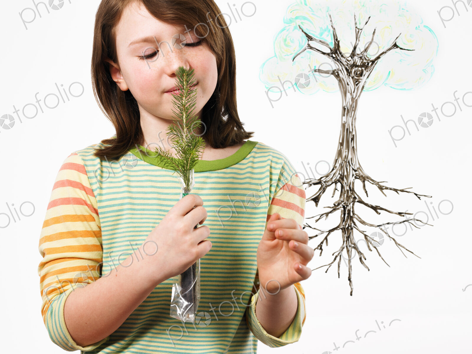 A young girl holding a small evergreen seedling to her nose and smelling it. An…