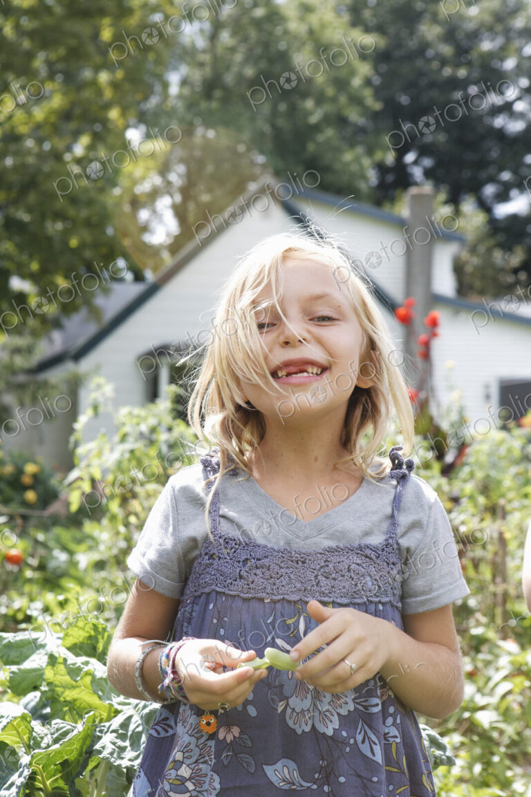 A young girl in a blue teeshirt and pinafore dress laughing, and holding green beans…