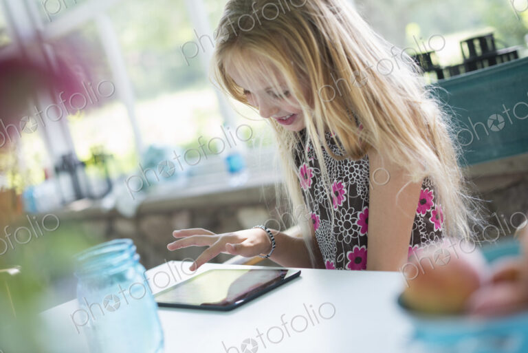 A young girl in a flowered dress using a digital tablet. Sitting at a table.