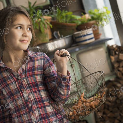 A young girl in a house kitchen, holding a metalwork basket of fresh carrots. A young girl in a house kitchen, holding a metalwork basket of fresh carrots.