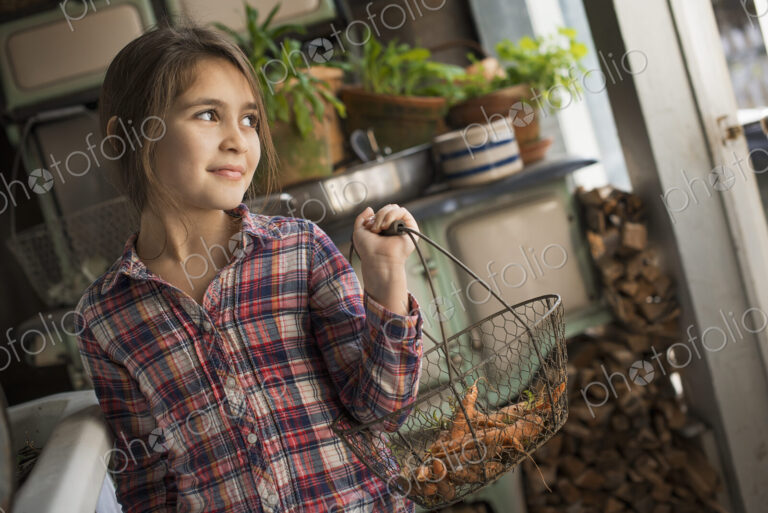 A young girl in a house kitchen, holding a metalwork basket of fresh carrots.