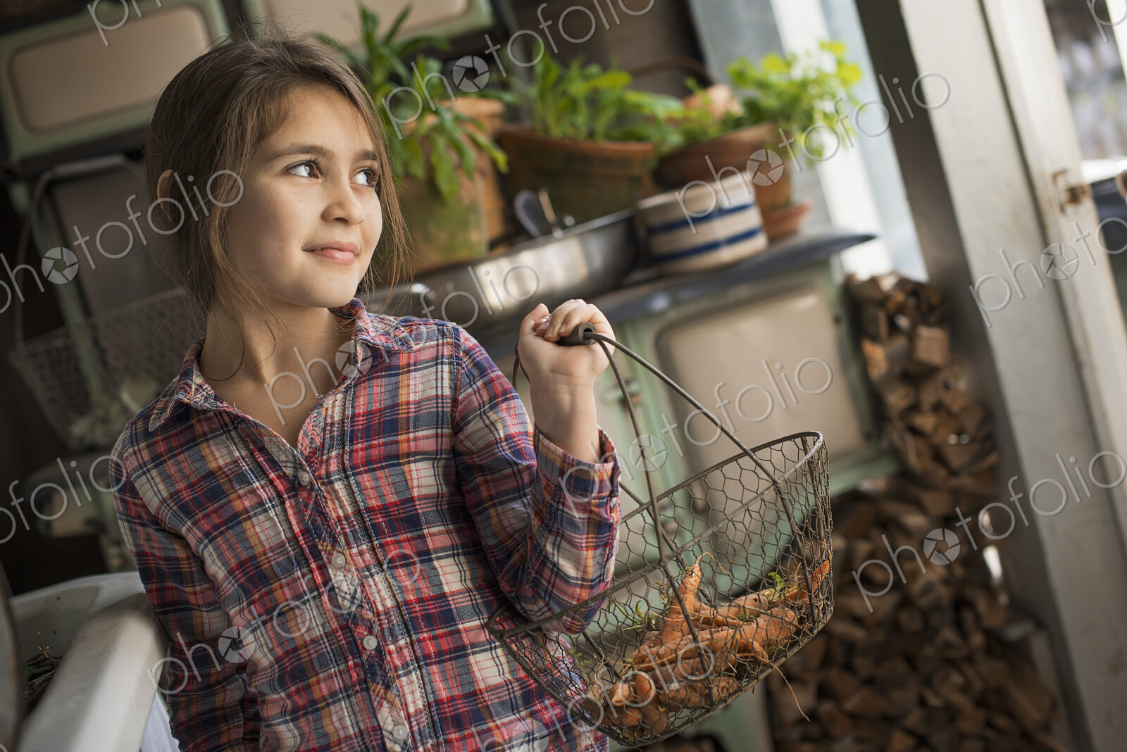 A young girl in a house kitchen, holding a metalwork basket of fresh carrots.