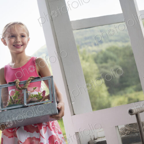 A young girl in a kitchen wearing a pink dress. Carryng a terarium containing small…