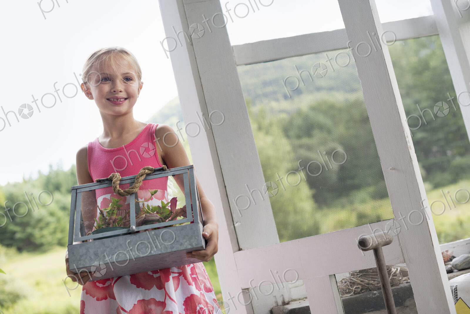 A young girl in a kitchen wearing a pink dress. Carryng a terarium containing small…