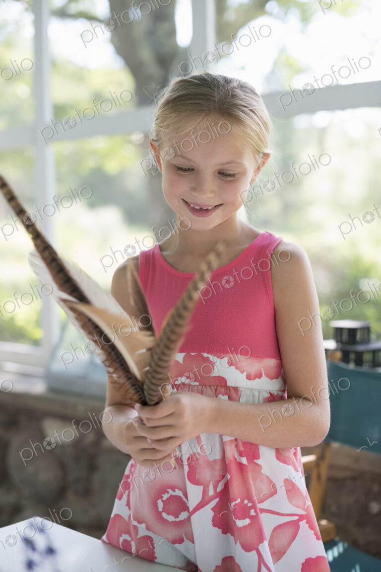 A young girl in a kitchen wearing a pink dress. Holding a bunch of bird feathers.