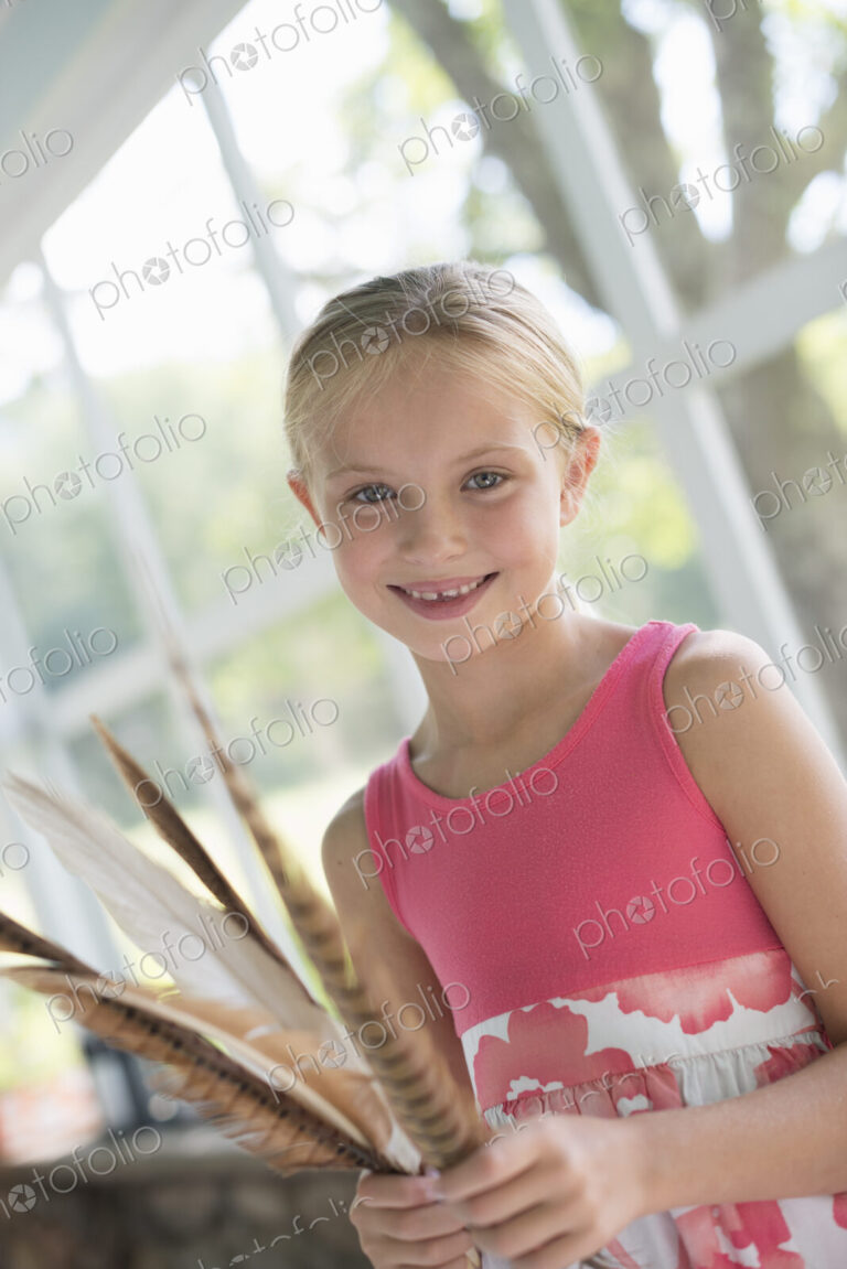 A young girl in a kitchen wearing a pink dress. Holding a bunch of bird feathers.