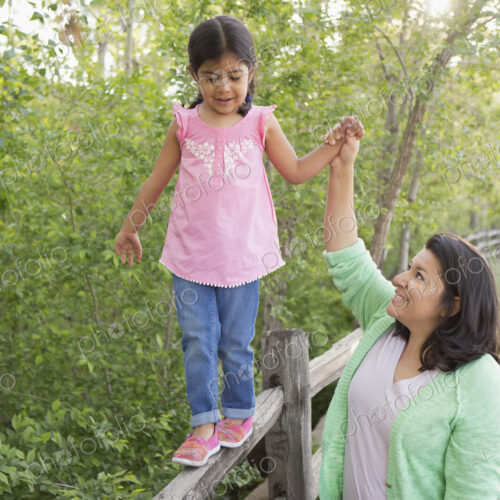 A young girl in a pink shirt and jeans, walking along a fence holding her mother's hand.