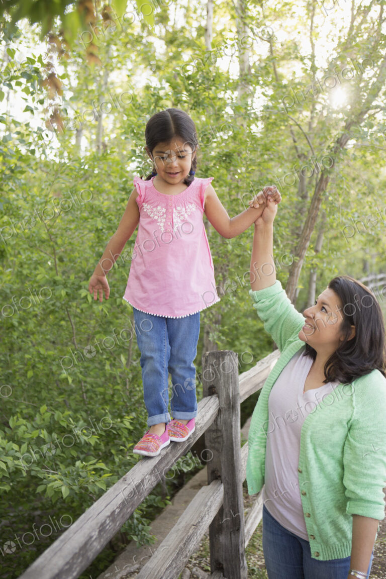 A young girl in a pink shirt and jeans, walking along a fence holding her mother's hand.
