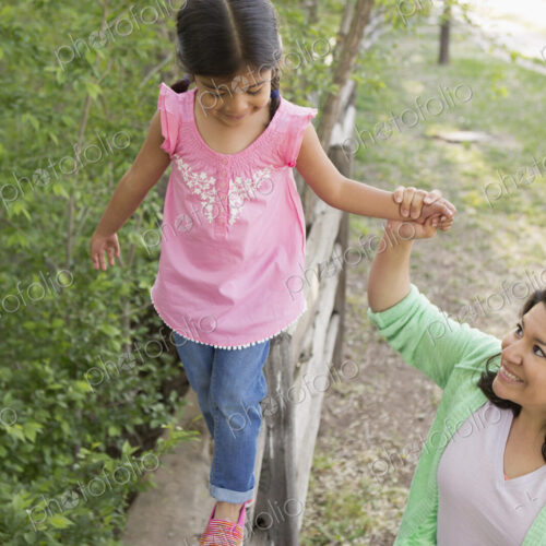 A young girl in a pink shirt and jeans, walking along a fence holding her mother's hand.