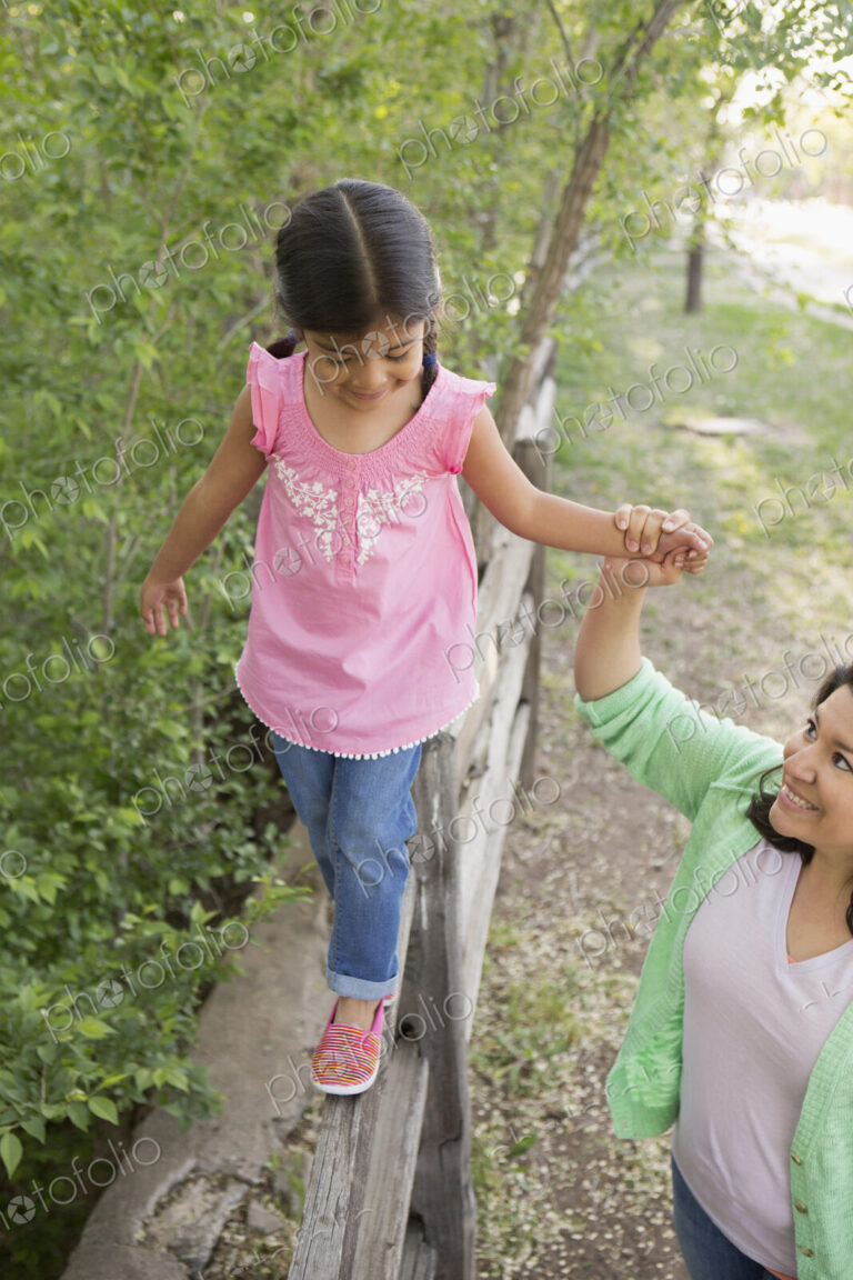 A young girl in a pink shirt and jeans, walking along a fence holding her mother's hand.