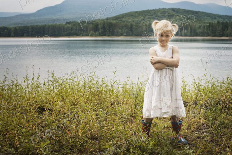 A young girl in a white summer dress, standing with her arms folded looking at the camera.