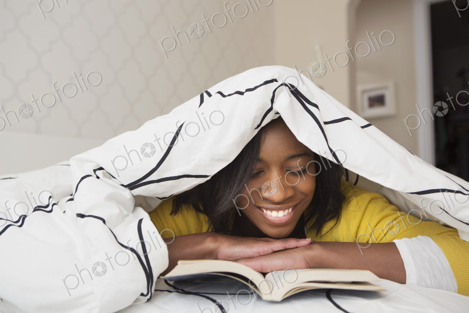 A young girl in her bed, under a duvet reading a book.