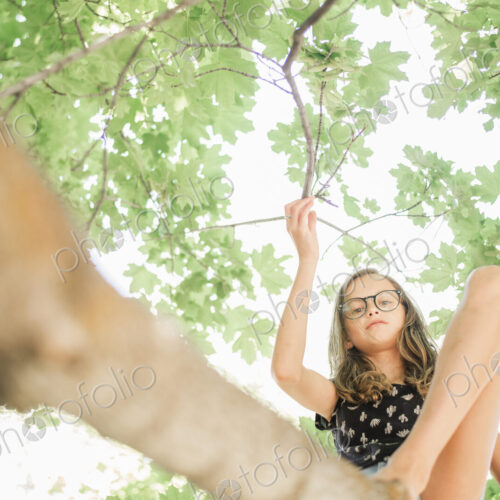 A young girl in shorts sitting on a high tree branch under a canopy of green leaves. A young girl in shorts sitting on a high tree branch under a canopy of green leaves.