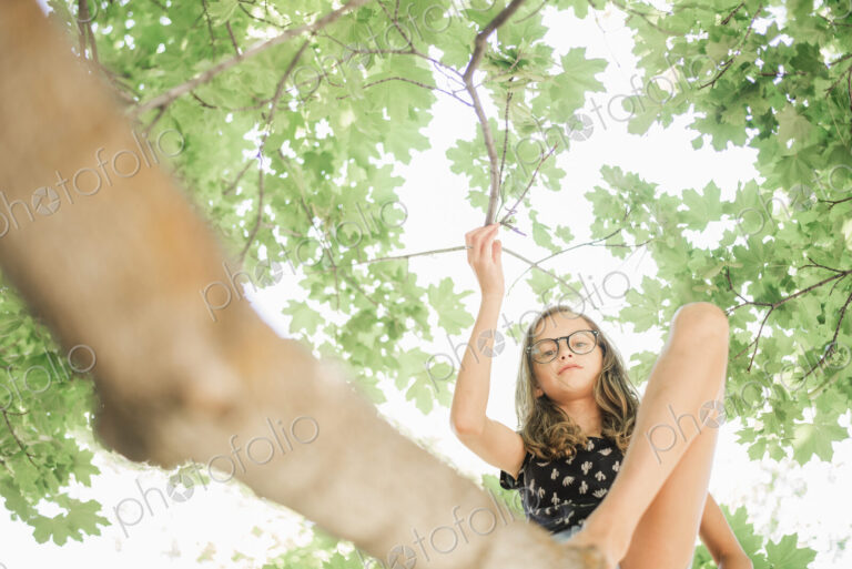 A young girl in shorts sitting on a high tree branch under a canopy of green leaves.