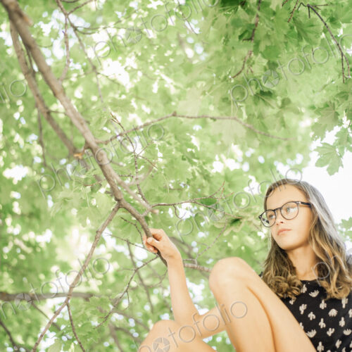 A young girl in shorts sitting on a high tree branch under a canopy of green leaves. A young girl in shorts sitting on a high tree branch under a canopy of green leaves.