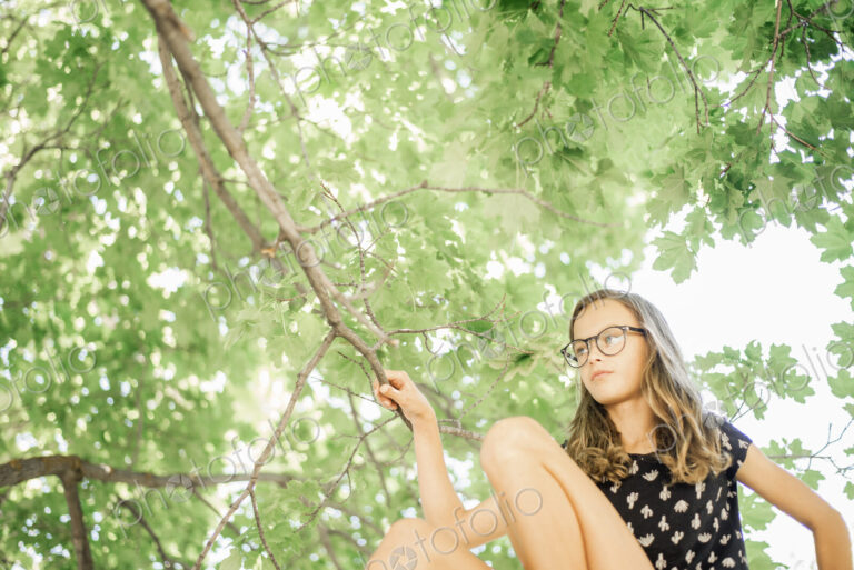 A young girl in shorts sitting on a high tree branch under a canopy of green leaves.