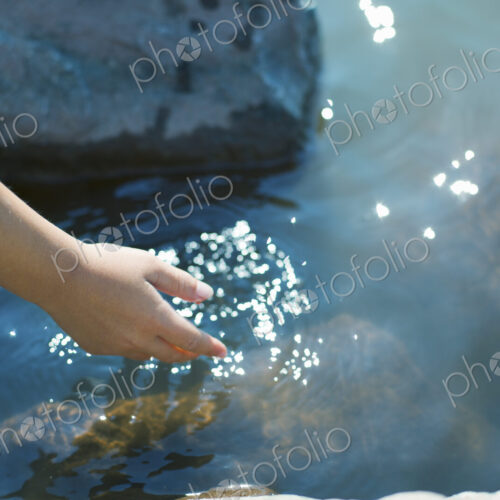 A young girl leaning forward to put her hand into clear shallow lake water.