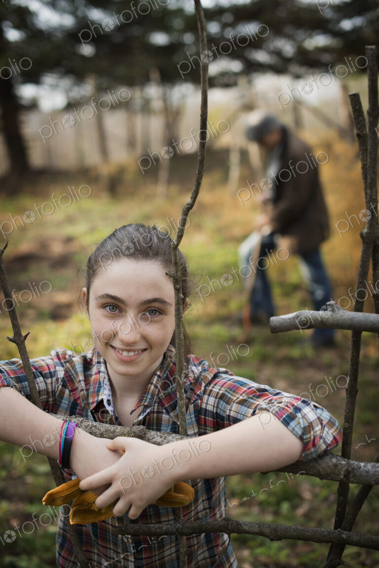 A young girl leaning on a fence, on an organic farm. A man digging the soil in the…