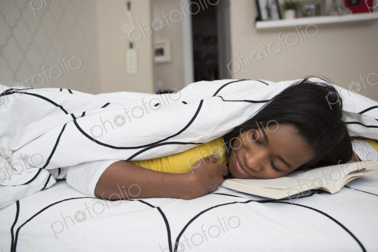 A young girl lying in her bed with her face resting on an open bok.