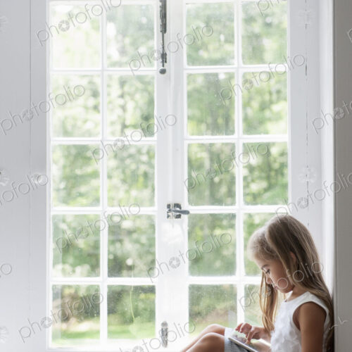 A young girl sitting at a window seat, using a digital tablet.