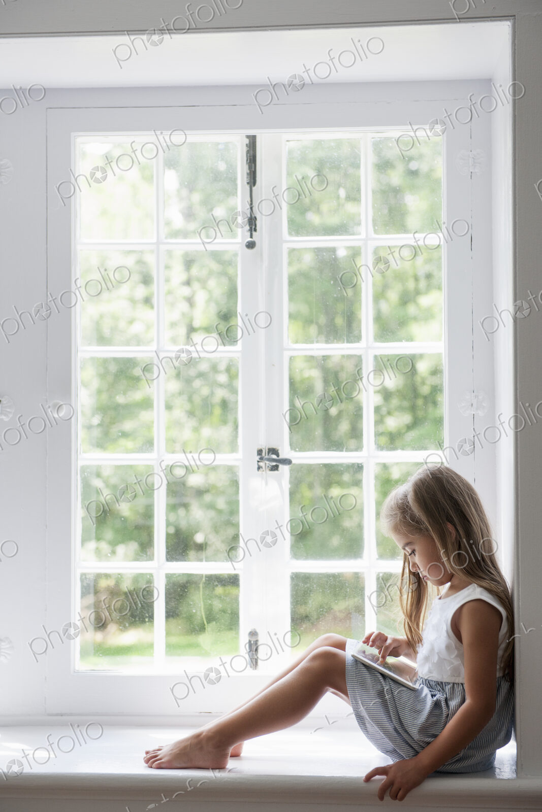 A young girl sitting at a window seat, using a digital tablet.