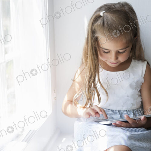 A young girl sitting at a window seat, using a digital tablet.
