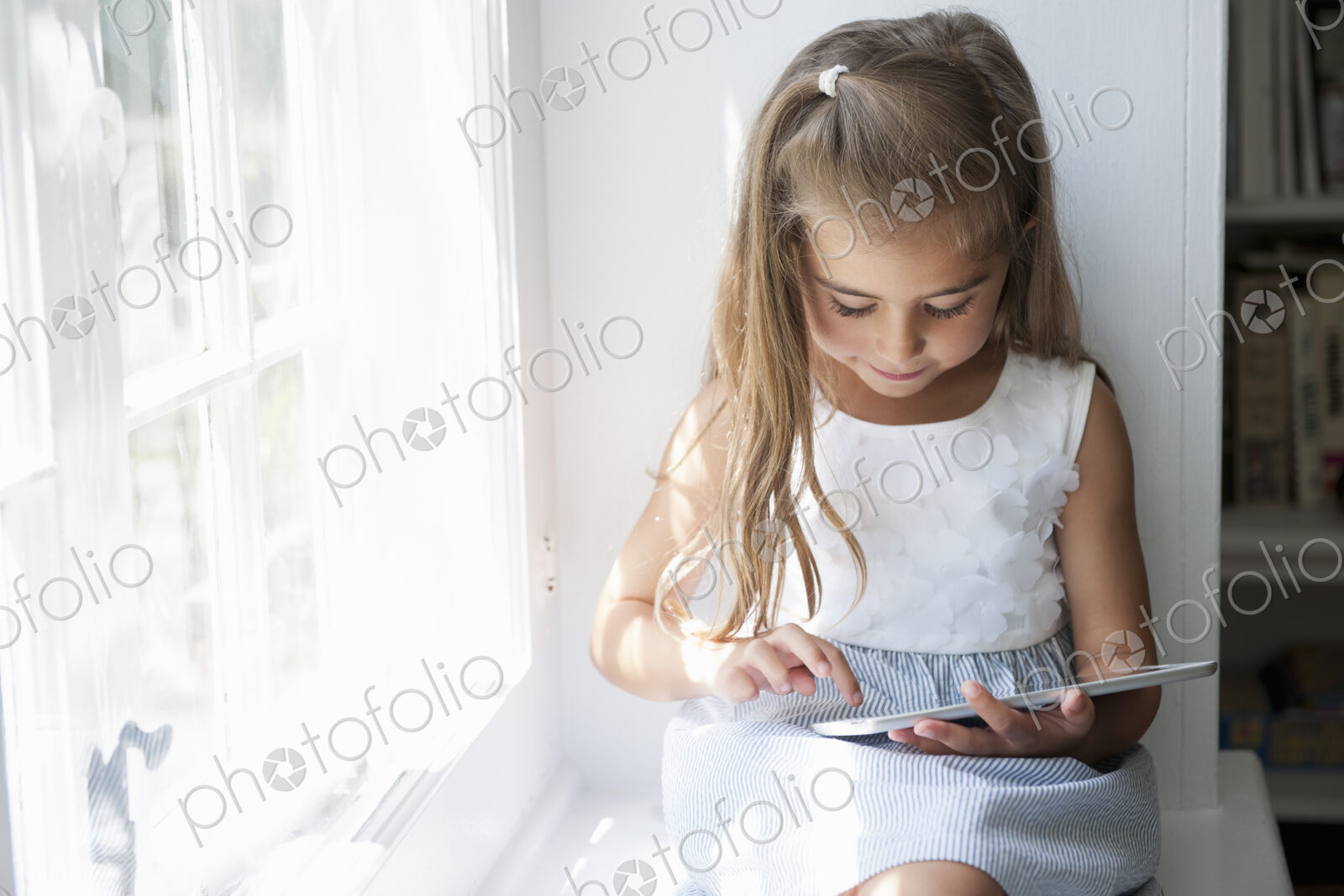 A young girl sitting at a window seat, using a digital tablet.