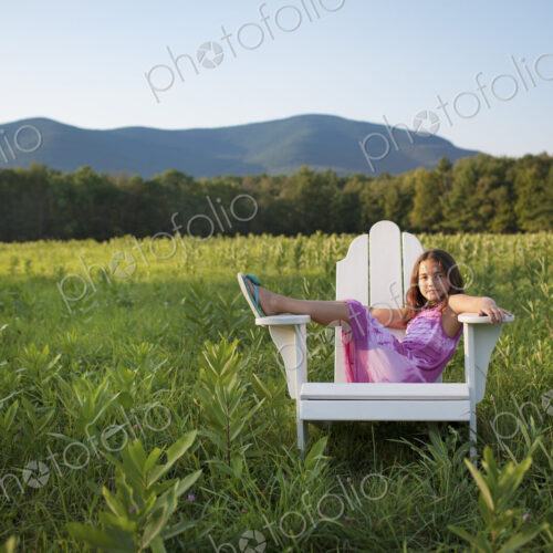 A young girl sitting in a traditional wooden Adirondack style chair in a field at…