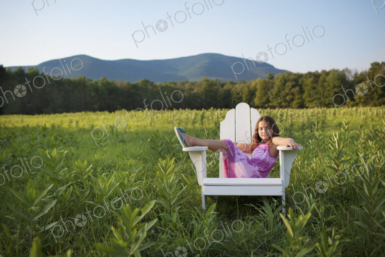 A young girl sitting in a traditional wooden Adirondack style chair in a field at…