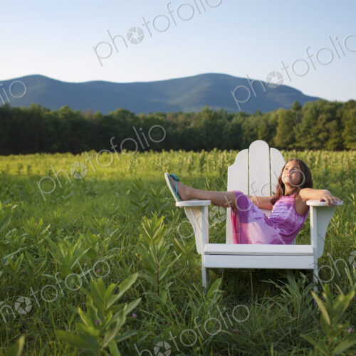 A young girl sitting in a traditional wooden Adirondack style chair in a field at…