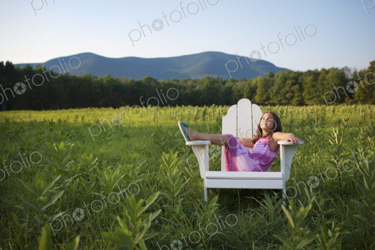 A young girl sitting in a traditional wooden Adirondack style chair in a field at…