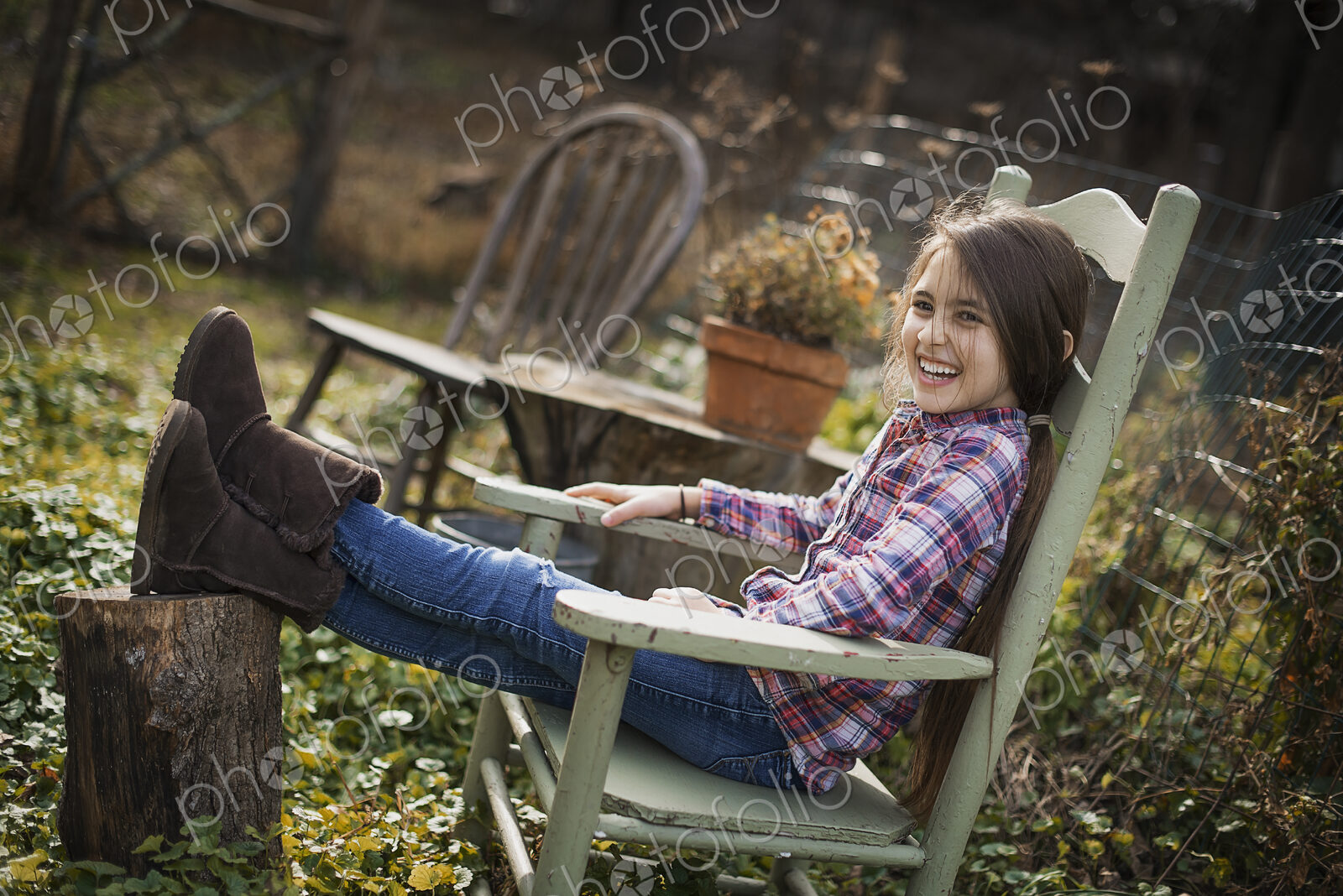 A young girl sitting in a wooden chair in a garden, with her feet up on a log.