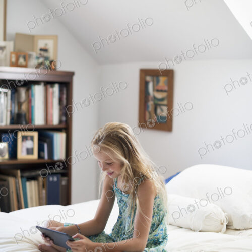 A young girl sitting on her bed using a digital tablet.