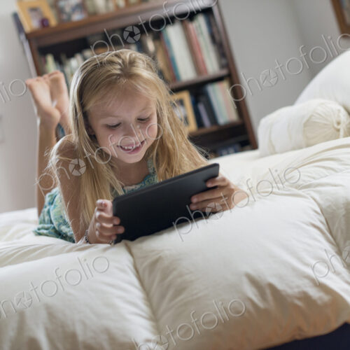 A young girl sitting on her bed using a digital tablet.