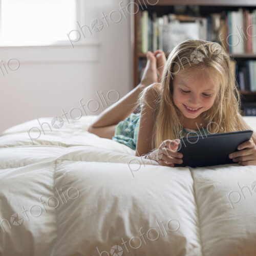 A young girl sitting on her bed using a digital tablet.