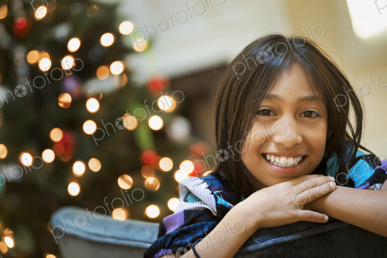 A young girl smiling and leaning over a chairback, in front of a Christmas tree.