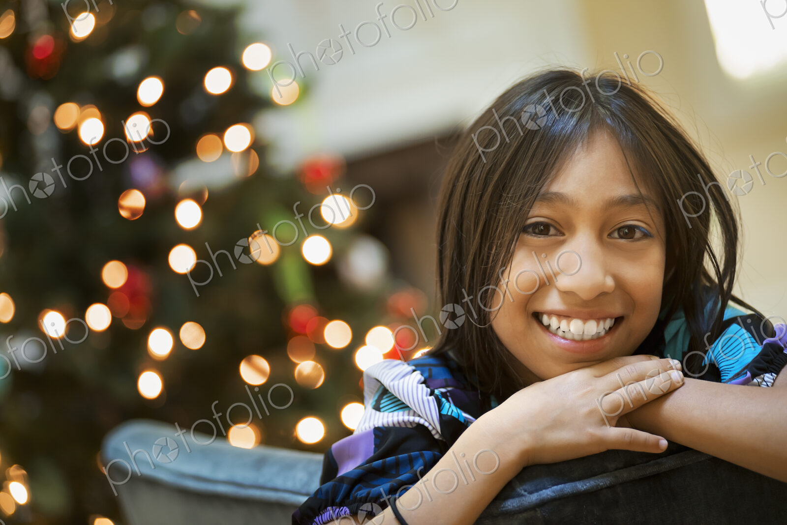 A young girl smiling and leaning over a chairback, in front of a Christmas tree.