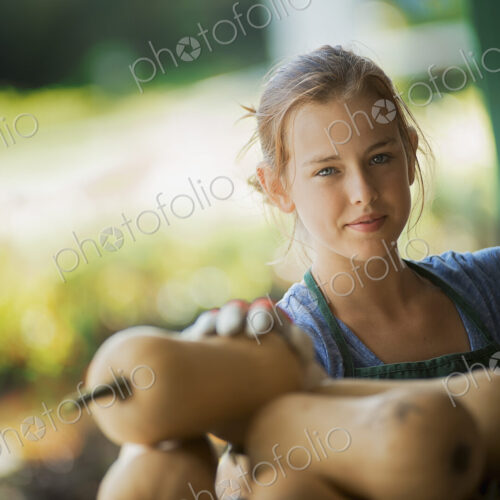 A young girl wearing a green apron, carrying a box of freshly picked butternut squash…
