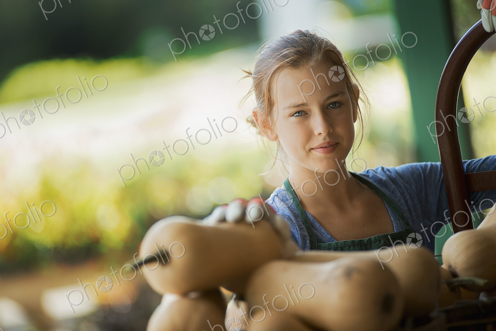 A young girl wearing a green apron, carrying a box of freshly picked butternut squash…